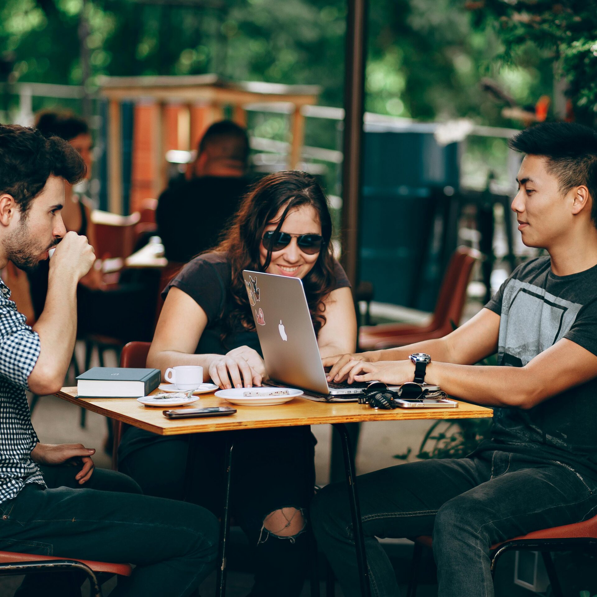 A group of young adults working on a laptop at an outdoor coffee shop, enjoying teamwork and collaboration.